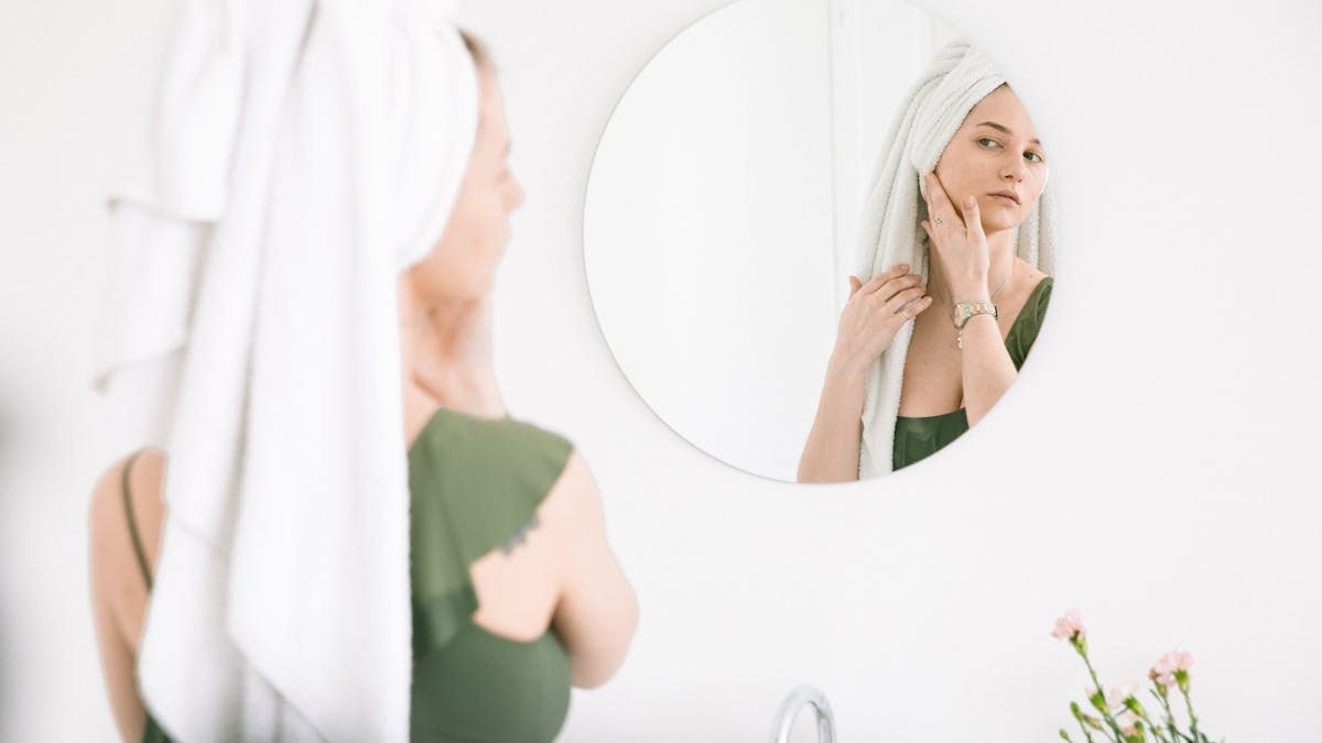 Woman in bathroom checking skin in mirror with towel on head.